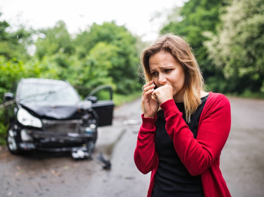 Frustrated woman calling for help on smartphone beside damaged car after accident